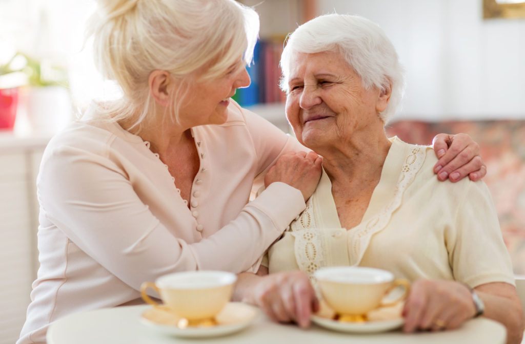 A senior woman and her daughter sitting on a couch smiling and talking to each other while holding a cup of tea.