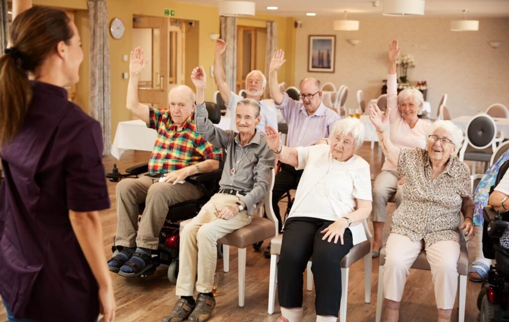 A group of seniors sit in chairs and raise their arms above their heads during a group exercise class at their assisted living community.