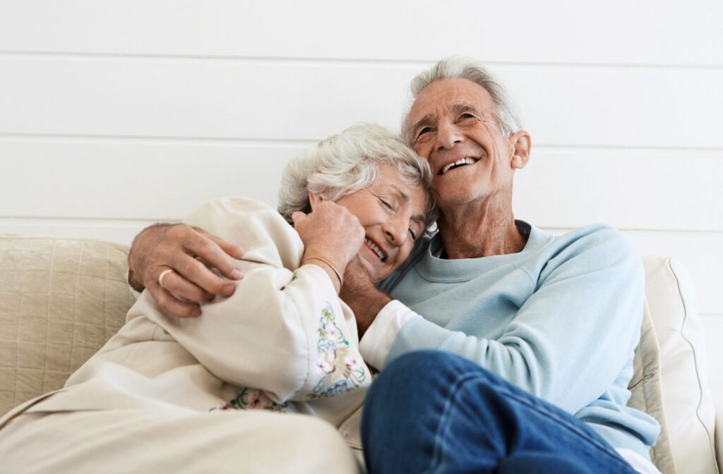 A senior couple hugs each other and leans back onto their cream colored coach. They are smiling and look content.