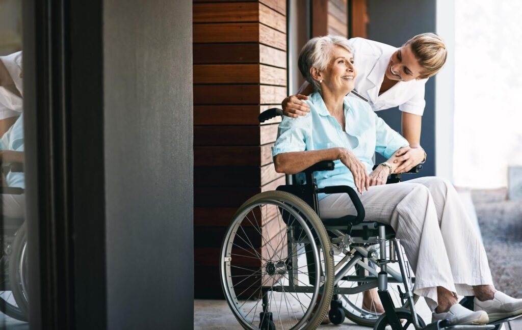 A fully-trained caregiver leans down to speak to a resident in a wheelchair while smiling.