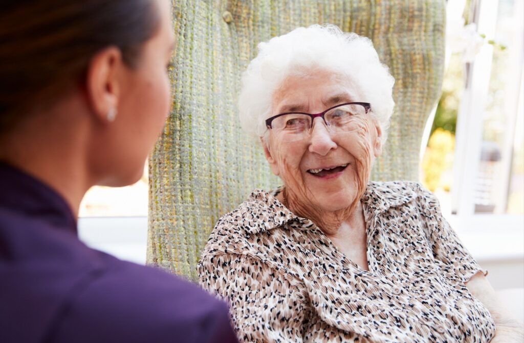 An older adult sits in a high-backed chair and smiles at a kneeling caregiver during a check-in